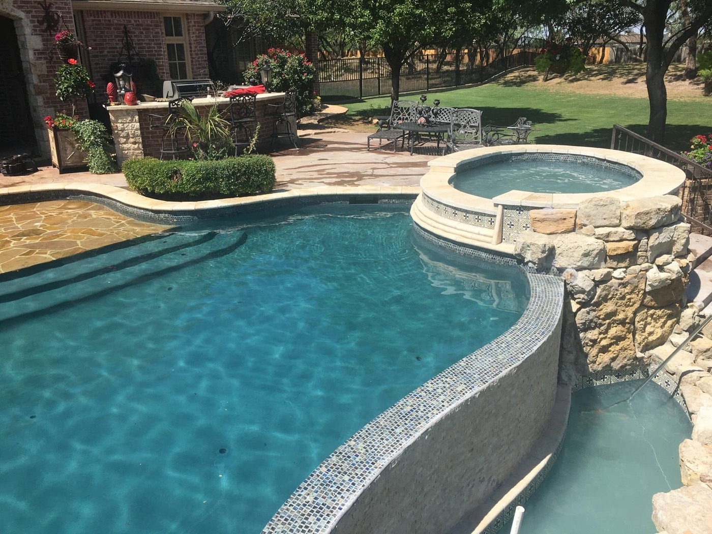 Finished freeform pool with raised tiled spa, natural stone, and outdoor kitchen